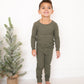 Child wearing green pajamas standing next to a small Christmas tree on a white floor.