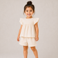 A young child wearing a cream-colored two-piece outfit with a ruffled short-sleeve top and matching shorts, paired with beige shoes, standing against a neutral light gray studio background.