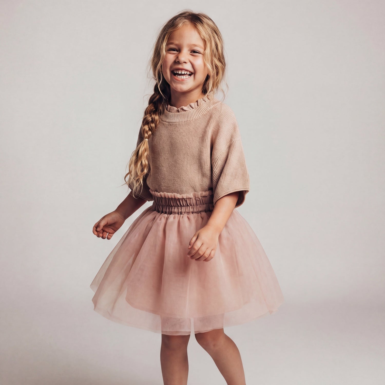 Young girl wearing a pinkish-tan sweater and pink tulle skirt on a plain background